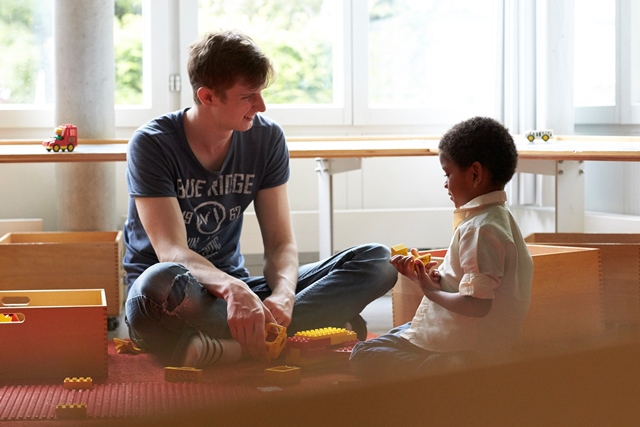 Ein Lernender Fachmann Kinderbetreuung EFZ sitzt mit einem Kind am Boden und baut Lego.
